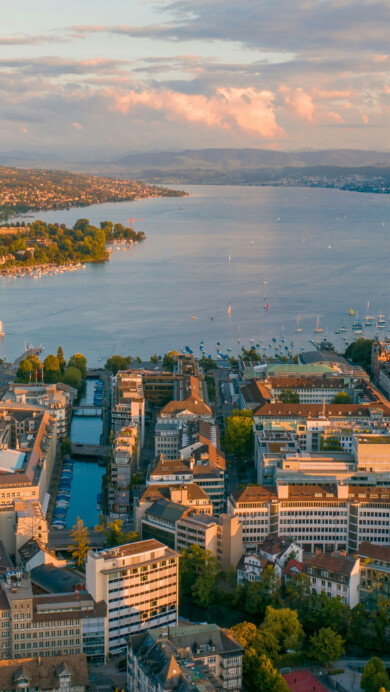 A mesmerizing 4K wallpaper captures the enchanting beauty of Zurich at sunset from an aerial perspective. The city's skyline is illuminated by the warm hues of the setting sun, casting a golden glow over the urban landscape. Lights from buildings twinkle as dusk settles in, creating a picturesque scene ideal for adorning your desktop or mobile screen.