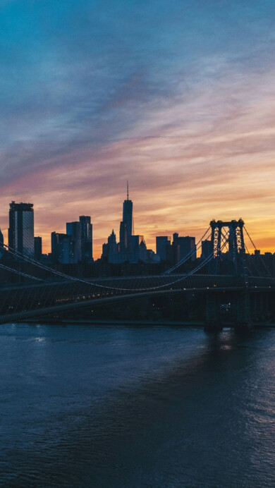 A stunning 4K wallpaper featuring the Williamsburg Bridge at sunset, with the New York skyline silhouetted against a vibrant evening sky. The warm hues of the sunset reflect off the water, creating a picturesque and serene scene perfect for your desktop or mobile background.