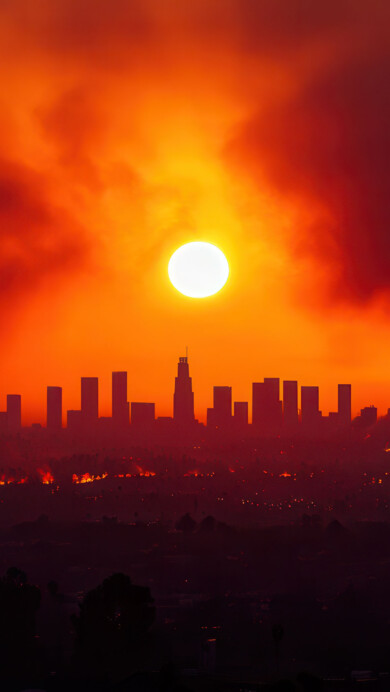 A stunning 4K wallpaper featuring a dramatic firestorm sweeping over the skyline of Los Angeles. The fiery orange and red hues of the wildfire contrast with the darkening sky, creating a powerful visual scene that captures the intensity of the moment, perfect for your desktop or mobile wallpaper.